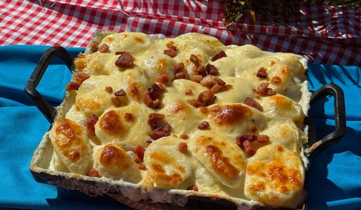 Fotografia de uma panela quadrada preta de ferro sobre um pano de mesa azul. Dentro da panela tem uma receita de batata gratinada com molho de requeijão, queijo e bacon.