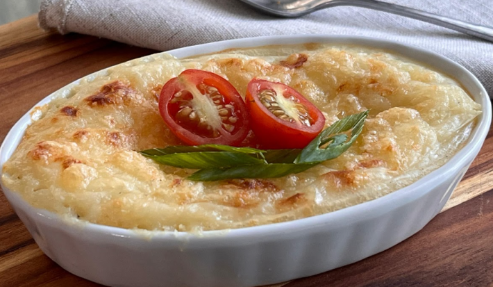 A imagem mostra uma travessa oval branca com um purê de batata gratinado em tons de dourado e amarelo.