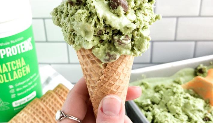 Fotografia em tons de verde em uma bancada de madeira, com um fundo de azulejo, com uma pessoa segurando uma casquinha com o sorvete de matcha, um pote de produto de Vital Proteins. E ao fundo, um recipiente com mais sorvete.