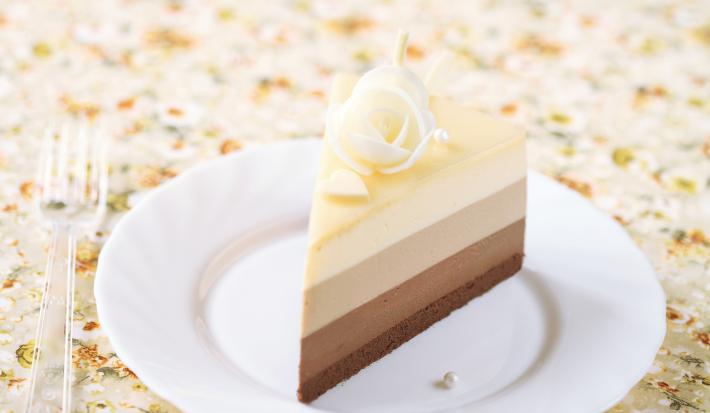 Fotografia de tons claros de uma torta diplomata, com creme preto e branco em camadas em um prato branco de sobremesa. Ao lado, um garfo pequeno de plástico sobre uma toalha de mesa colorida.