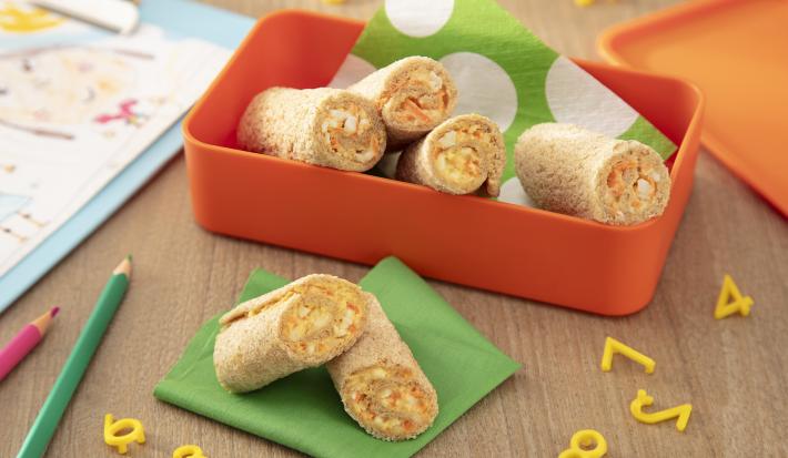 Fotografia de um recipiente laranja quadrado e fundo com enroladinhos de pão com cenoura e ovo. Dentro do recipiente tem um papel verde e branco, e ao lado tem mais dois rolinhos sobre um papel verde dobrado e uma mesa de madeira.