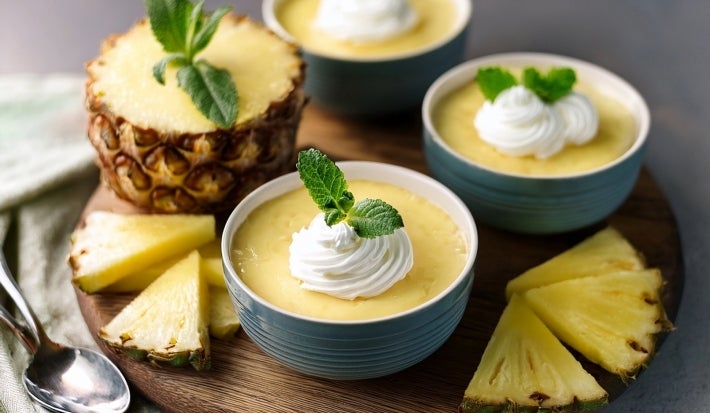 Fotografia de tigelas azuis, com creme amarelo, cobertura de chantilly e folhas de hortelã. Um dos doces é servido em uma metade de abacaxi, rodeado por fatias de abacaxi fresco em uma tábua de madeira.