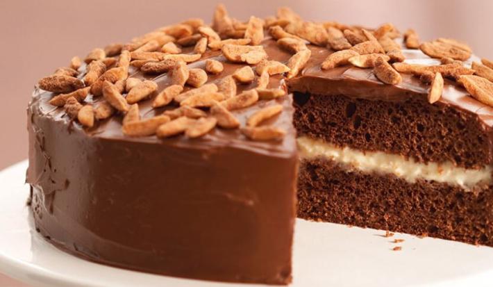 Fotografia em tons de rosa em uma mesa com uma toalha bege, um guardanapo de pano rosinha, um suporte para bolo branco com o bolo de chocolate cortado ao meio em cima dele.