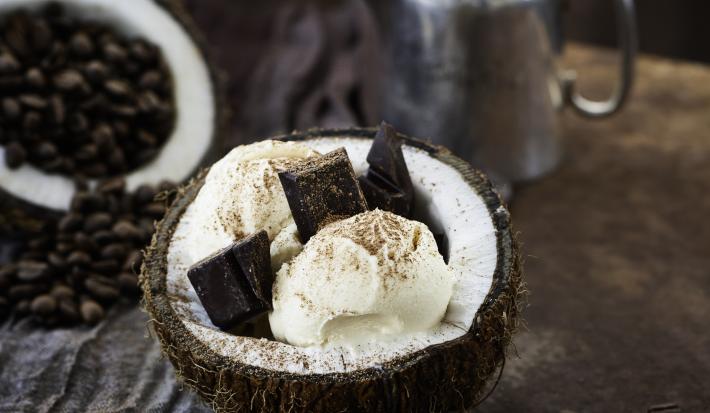 Fotografia de duas bolas de sorvete de coco com pedaços de chocolate dentro de um coco aberto, polvilhada com cacau em pó. O fruto está sobre uma mesa de madeira. Ao redor, cacau e nuts em potes pequenos na cor azul.