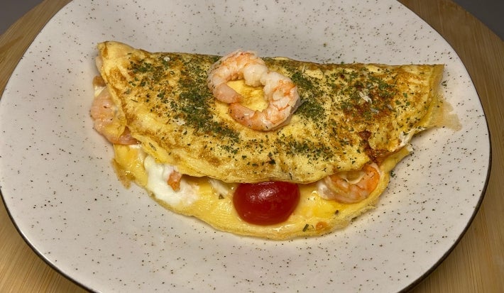 Fotografia de uma omelete dobrado ao meio com recheio cremoso de camarão e tomates cereja. Por cima da omelete tem dois camarões pequenos, e a omelete está sobre um prato raso de vidro branco.