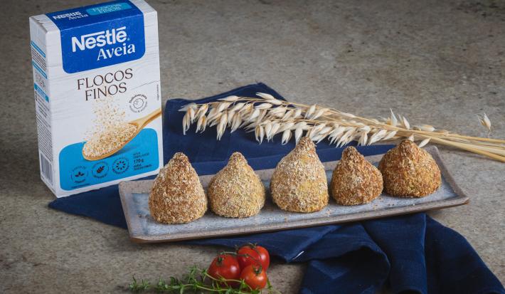 Foto da Receita de Coxinha da Batata Doce com Nestlé Aveia Flocos Finos, servidas em um prato cinza retangular, ao todo são 5, numa bancada de pedra com um tecido azul escuro, uma caixa de nestlé aveia flocos finos.