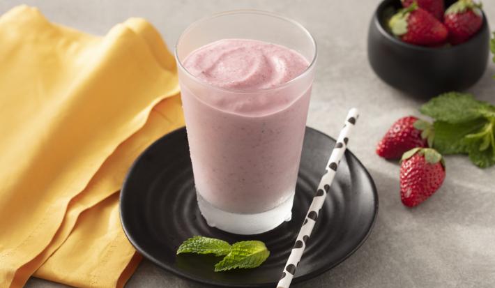 Fotografia em tons de amarelo, branco e preto de uma bancada branca com um prato preto redondo, sobre ele um copo de vidro com smoothie de morango e um canudo branco com bolinhas pretas. Ao lado um paninho laranja e morangos.