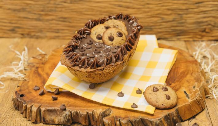 Fotografia em tons de marrom de uma bancada marrom com uma tábua de madeira com um paninho amarelo e branco, sobre ele um ovo recheado de cookie. Ao lado um cookie.