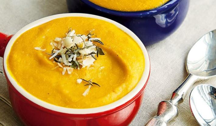 Fotografia em tons de vermelho e amarelo em uma mesa com uma toalha cinza, e duas panelinhas pequenas, vermelha e azul, com o creme de abóbora dentro delas.