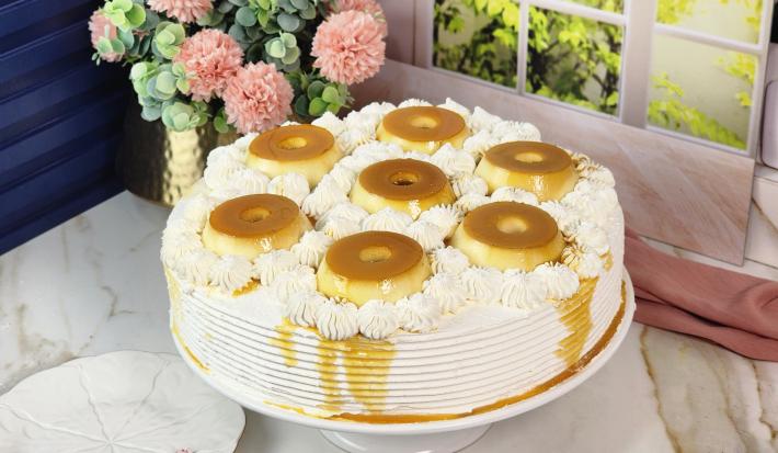 Foto da Receita de Bolo Recheado de Pudim com Doce de Leite Moça, decorado sobre um prato branco com sete mini pudins por cima, em uma bancada de mármore com flores ao fundo