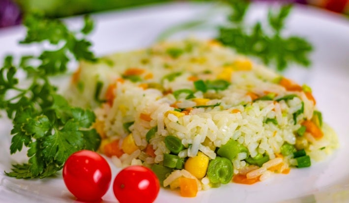 Imagem de frente de um prato de porcelana branco com uma porção colorida de arroz amarelo, milho verde,  ervilha, decorados com dois tomates-cerejas e galhos de coentro.