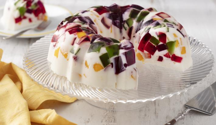 Fotografia em tons de branco e amarelo de uma bancada branca, sobre ele um prato de vidro com a gelatina mosaico. Ao fundo um prato redondo com uma fatia da gelatina. Ao lado um paninho amarelo.