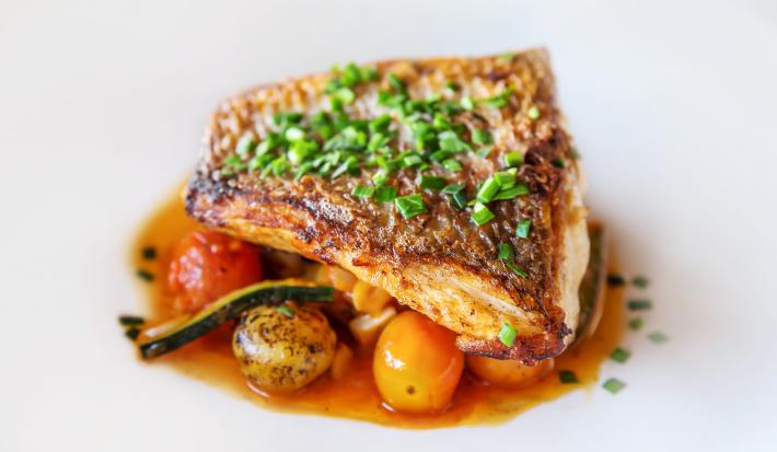 Fotografia de um filé de pescado ao papillote com alguns legumes ao forno por baixo do peixe. Por cima do peixe, que está em um prato branco, tem cebolinha salpicada.