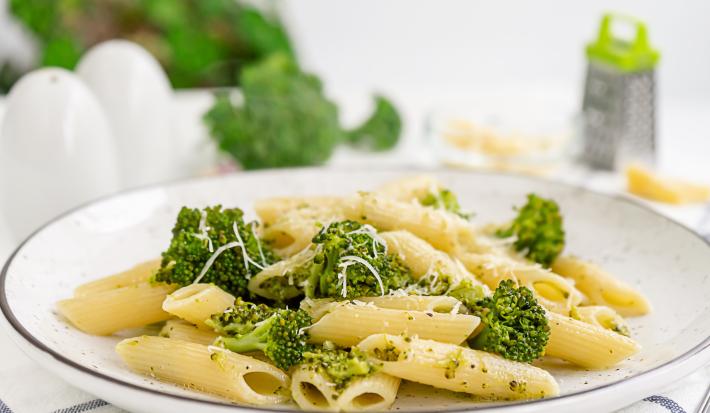 fotografia em tons de branco, bege e verde vista de frente. Contém um prato redondo branco com macarrão tipo penne e brócolis