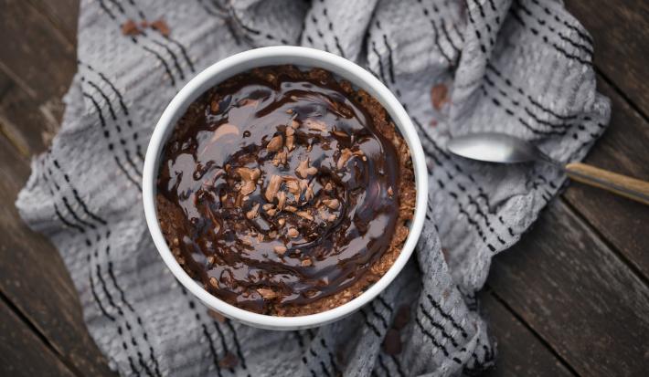 Fotografia vista de cima de um baked oats de air fryer dentro de um ramekin branco apoiado em um pano de tom cinza amassado, o qual está sobre uma madeira escura. Sobre o bolo tem uma calda de chocolate.