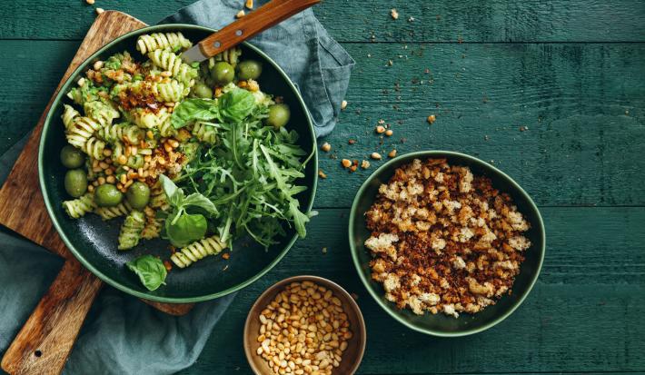 Fotografia vista de cima de uma salada verde com macarrão parafuso com iogurte, azeitona e nozes sobre uma tábua de madeira quadrada. Ao lado, dois bowls com grãos e croutons. A tábua está apoiada em um pano azul, que está sobre uma mesa em tom verde.