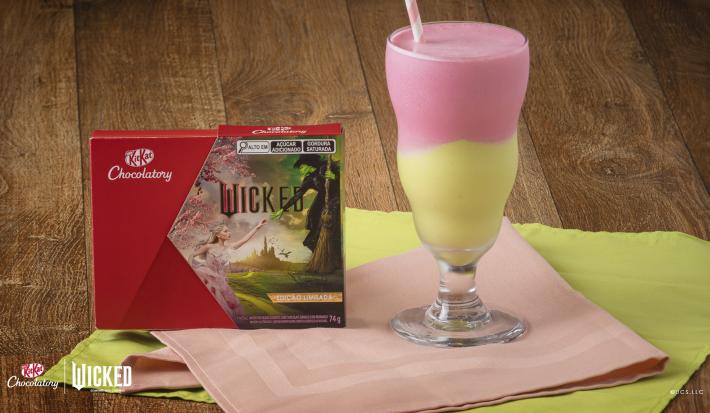Foto aproximada de um copo alto com a receita de milkshake dividido em cores verde e rosa, servido sobre uma bancada de madeira com tecidos verde e rosa e com a caixa de KitKat Wicked ao lado esquerdo