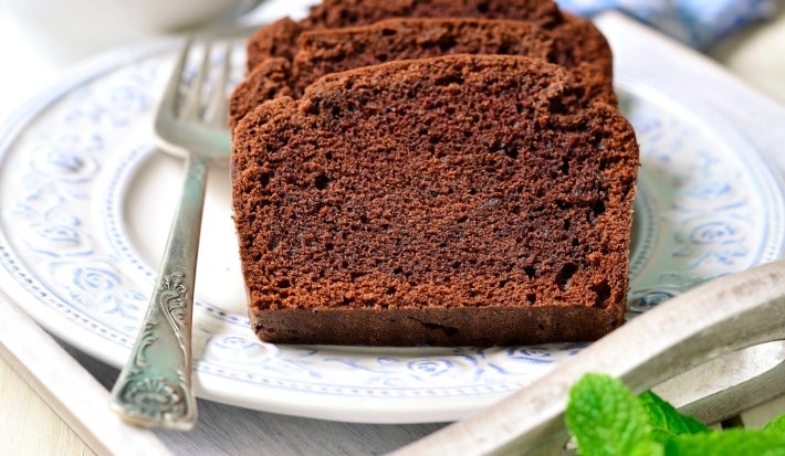 Fatias de bolo de chocolate fofinho em um prato decorado com flores azuis. Um garfo prateado ao lado e uma folha de hortelã adicionam charme. O fundo desfocado com um pano xadrez cria uma atmosfera acolhedora.