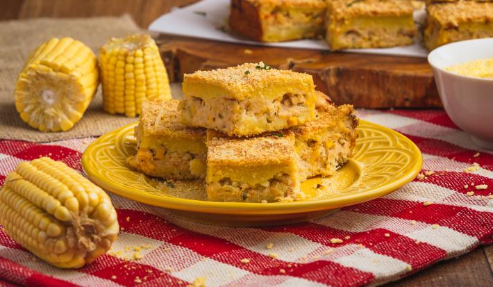 Foto de uma bancada de madeira com uma toalha vermelha quadriculada, com um prato amarelo no centro e pedaços da torta de milho com frango. Há milhos decorando a cena e, ao fundo, mais pedaços de torta sobre uma tábua de madeira.
