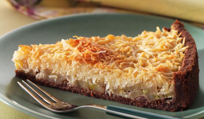 Fotografia em tons de amarelo em uma bancada de madeira, um pano amarelo com flores coloridas, um prato verde redondo raso com uma fatia da torta de coco com banana.