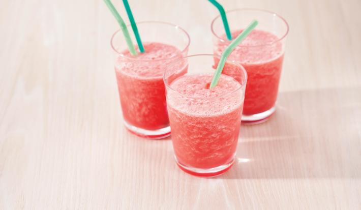 Fotografia em tons de rosa em uma bancada de madeira clara com três copos de vidro com o smoothie de melancia dentro deles e canudinhos verdes dentro dos copos.