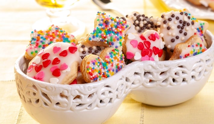 Em cima de uma mesa, tigelas com biscoitos redondos e em formato de coração com glacê branco e confeitos coloridos.