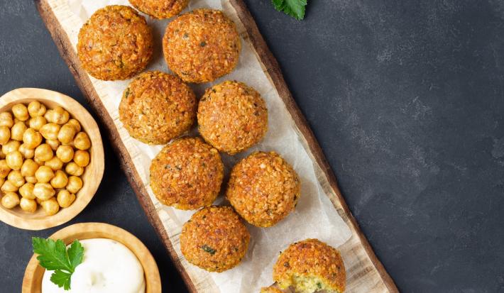 Fotografia vista de cima de oito unidades de falafel sobre um apoio de madeira claro retangular, ao lado de um falafel aberto também sobre o apoio. Ao lado do apoio tem dois recipientes de madeira com grão de bico e um molho de iogurte.