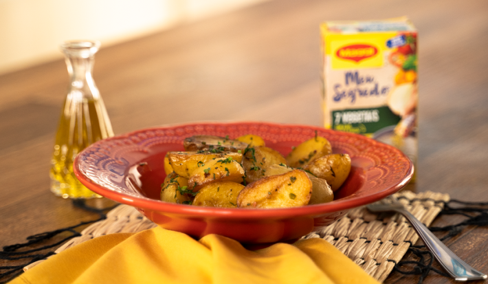 Foto da Receita de Batata Bolinha Sautée. Observa-se um prato vermelho com as batatinhas. Do lado esquerdo, um pote transparente com azeite e, do direito, um garfo e uma embalagem de Maggi Meu Segredo 7 Vegetais.