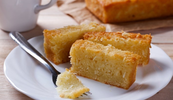 Três pedaços de bolo de coco estão em um prato branco, com um garfo e uma caneca ao fundo. Um pedaço foi cortado, revelando sua textura macia e úmida. A cena sugere um lanche caseiro simples e apetitoso.