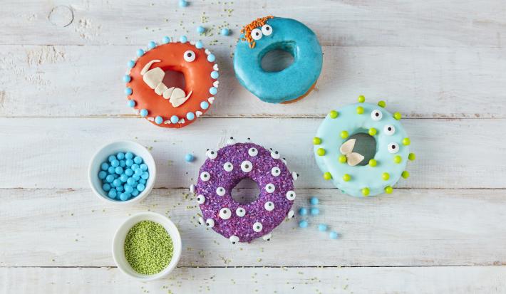 Imagem vista de cima de alguns Donuts Monstro em tons coloridos, decorados com tema de dia das bruxas, tudo em uma bancada de madeira em tom claro