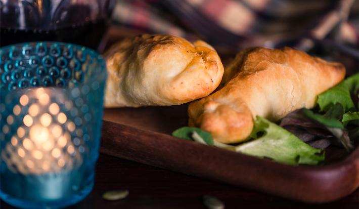 Fotografia em tons escuros duas empanadas recheadas com sardinha sobrepostas e apoiadas em uma tábua de madeira retangular escura. Na frente da tábua tem um copo de vidro com texturas de cor azul com uma vela dentro.