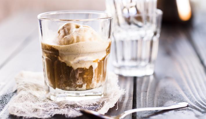 Fotografia de um copo de vidro com café, gelo e sorvete de creme que está em cima de um pano fino, ao lado de uma colher de sobremesa. Ao fundo, um copo de vidro vazio, sobre uma mesa de madeira escura.
