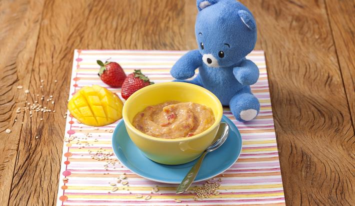 Fotografia em tons de amarelo em uma bancada de madeira e um paninho listrado colorido com um potinho amarelo com o creminho de manga com morango apoiado em um prato azul. Ao lado, um ursinho de pelúcia azul, dois morangos e uma fatia de manga.