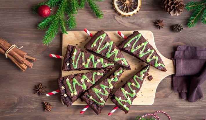 Em cima de uma mesa, tábua com brownie em forma de triângulo, decorado com creme verde e confeitos de bolinhas e estrelas.