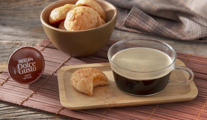 Foto da receita de pão de queijo 3 ingredientes servida em um bowl de cerâmica e à frente há uma xícara de café nescafé dolce gusto caffe matinal com um pão de queijo ao lado sobre uma tábua de madeira