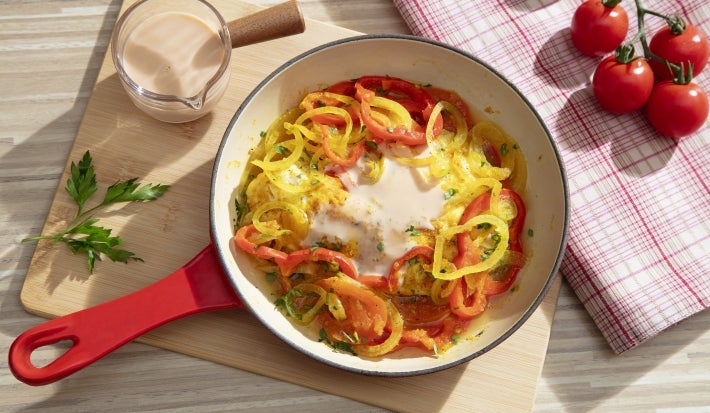 Fotografia em tons claros vista de cima de uma frigideira vermelha e branca com uma receita de moqueca e novasource. A frigideira está sobre uma tábau de madeira retangular grande ao lado de um pano brano com listras vermelhas e tomates pequenos.
