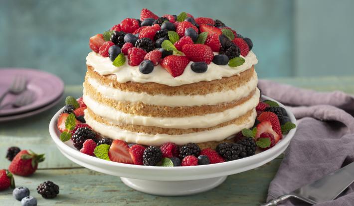 Fotografia em tons de vermelho e lilás em uma bancada de madeira verde, um pano roxo, um suporte para bolo com o bolo pelado (naked cake) com creme branco e com decoração de frutas vermelhas. Ao fundo, pratos pequenos lilás de sobremesa.