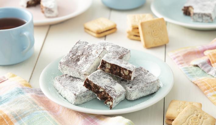 Foto em tons de azul claro da receita de palha italiana de passatempo com leite ninho servida em algumas porções sobre um prato de porcelana azul com biscoitos passatempo em volta além de uma xícara de café azul e um paninho quadriculado para decorar