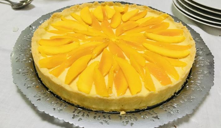 Fotografia em tons de amarelo em uma bancada de madeira de cor branca. Ao centro, uma mesa contendo a torta de manga. Ao fundo, alguns pratos brancos empilhados.