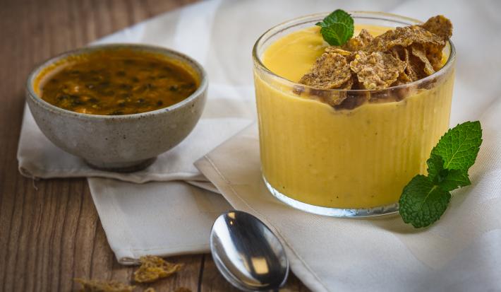 Fotografia em tons de amarelo em uma bancada de madeira, um paninho bege, um potinho com um creme de iogurte de maracujá com gengibre. Ao lado, um potinho de maracujá e uma colher.