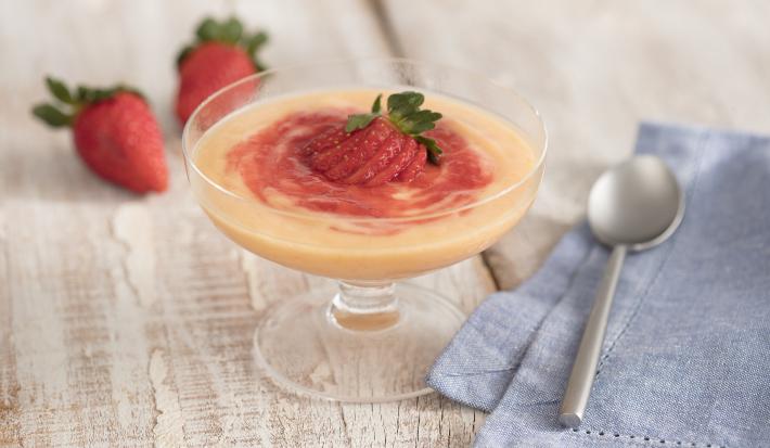 fotografia em tons de cinza e vermelho de uma bancada cinza vista de frente. Contém uma taça transparente com o creme dentro e um morango por cima para decorar, ao lado um pano azul e uma colher para servir e ao fundo dois morangos.
