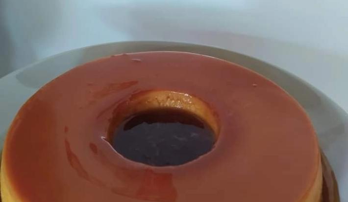 Fotografia em tons de caramelo, em uma bancada de madeira de cor branca. Ao centro, um prato redondo branco contendo o pudim.