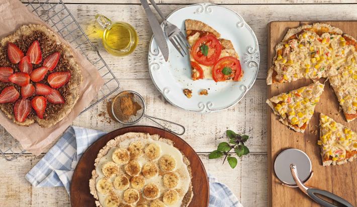 fotografia em tons de bege de uma bancada vista de cima, contém uma tábua redondo e marrom com uma pizza com fatias de banana, uma grade com uma pizza de chocolate e fatias de morango e um prato redondo e branco com uma fatia de pizza