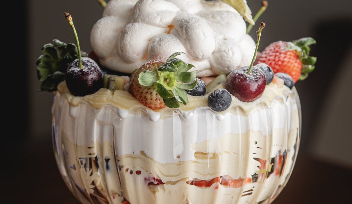 Foto aproximada da receita de Pavlova na taça, montada em uma linda taça de cristal com camadas de creme, merengue e frutas amarelas e vermelhas. Decorada com um lindo merengue no topo.