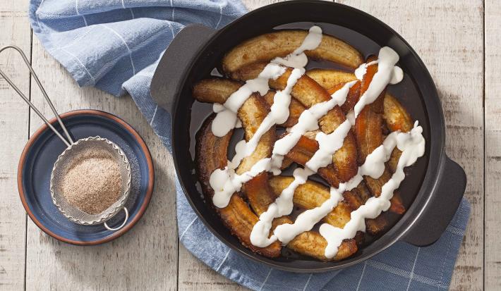 Fotografia em tons de dourado e branco, ao centro panela refratária com banana flambada e creme de leite, sobbre guardanapo azul com quadriculados brancos, ao lado potinho com uma peneirinha com açúcar e canela, sobre bancada de madeira rústica.
