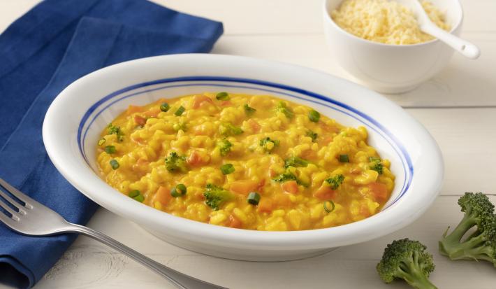 Fotografia em tons de azul em uma bancada de madeira clara com um pano azul escuro ao lado e um prato branco com detalhes em azul ao centro com o risoto de legumes com cúrcuma dentro. Ao lado, um potinho branco com queijo parmesão ralado e dois brócolis.