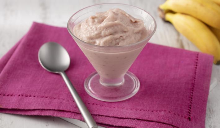 Fotografia em tons de branco e rosa de uma bancada branca com um paninho rosa, sobre ele uma colher e uma taça de vidro com o frozen. Ao fundo um cacho de banana.