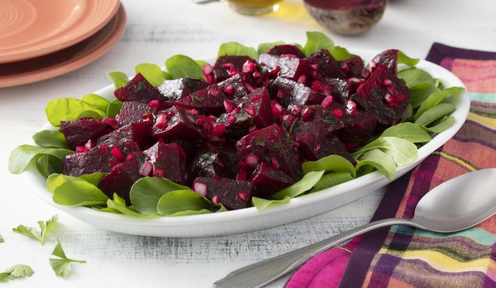 Fotografia em tons de roxo em uma bancada de madeira branca com um pano colorido xadrez com roxo, rosa e amarelo. Ao centro, um recipiente branco oval com salada de beterraba temperada com salsinha e cebola. Ao fundo, beterraba cortada ao meio.