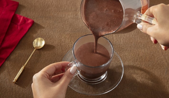 Fotografia mostra uma mão feminina segurando uma xícara de vidro sobre um prato pequeno, também de vidro. Dentro da xícara tem chocolate quente cremoso de nescau extra cacau que está caindo de um outro recipiente de vidro.