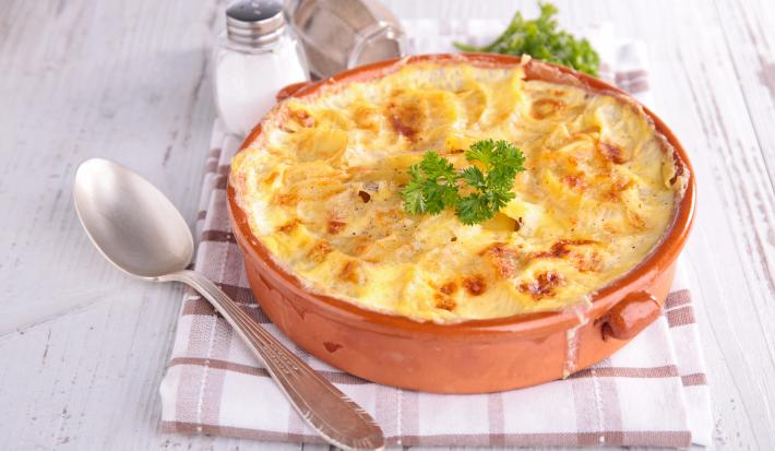 fotografia em tons de branco, vermelho e amarelo de uma bancada branca vista de cima. Contém um recipiente redondo vermelho com um gratinado de batatas, e ao lado uma colher para servir.